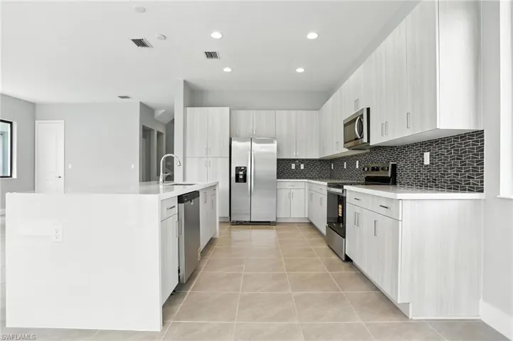 Kitchen featuring appliances with stainless steel finishes, light tile patterned floors, light stone countertops, decorative backsplash, and an island with sink