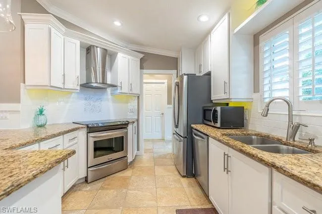 Kitchen with white cabinetry, backsplash, stainless steel appliances, wall chimney exhaust hood, and ornamental molding