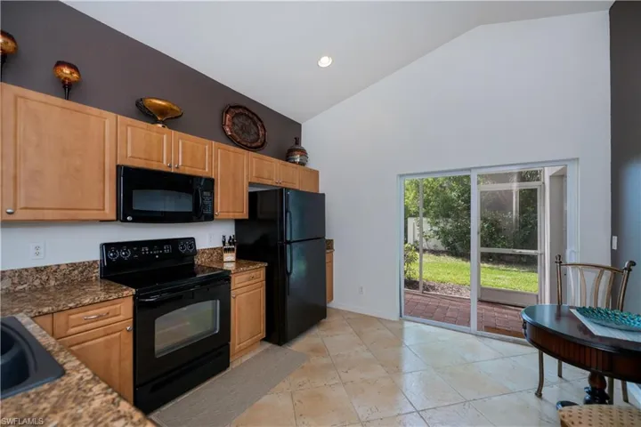 Kitchen with high vaulted ceiling, dark stone counters, black appliances, light brown cabinetry, and baseboards