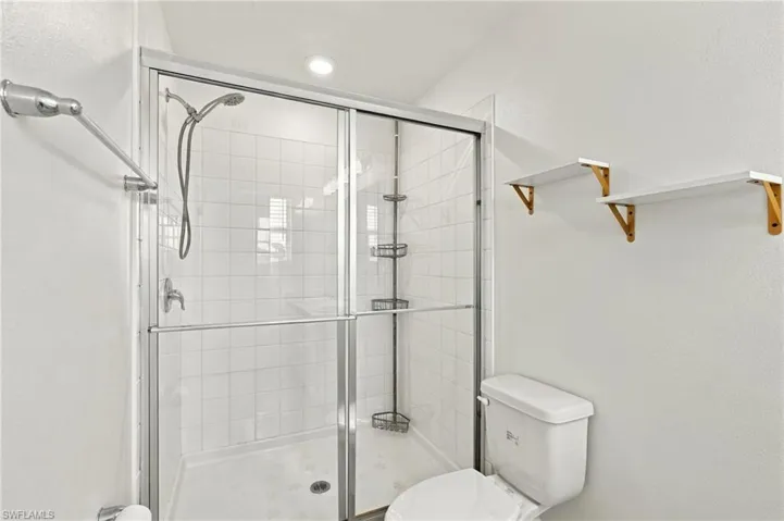Shower enclosure with white tile surround, sliding glass door, handheld shower fixture, built-in shelving, and white toilet