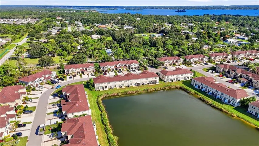 Aerial perspective of a multi-unit residential community featuring red-tiled roofing, paved street infrastructure, and a central pond, with a large body of water in the background