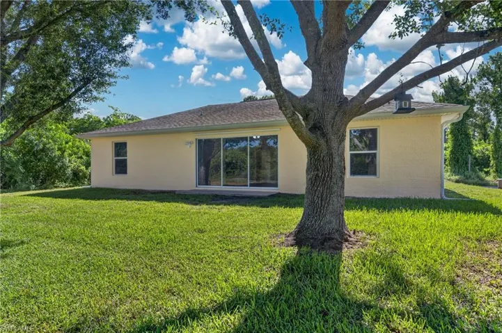 Expansive grassy yard featuring a mature tree, stucco exterior, multiple windows, and a sliding glass door for patio access