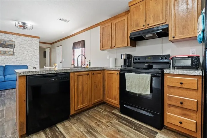 Kitchen featuring black appliances, a peninsula, under cabinet range hood, dark wood-style flooring, and brown cabinets
