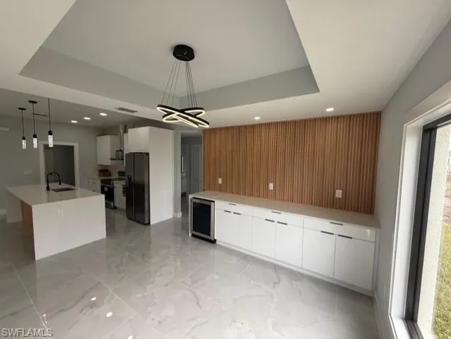 Kitchen featuring white cabinets, a tray ceiling, black fridge with ice dispenser, decorative light fixtures, and beverage cooler