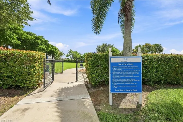 Paved pathway entrance to a fenced green space, featuring mature hedges and a palm tree
