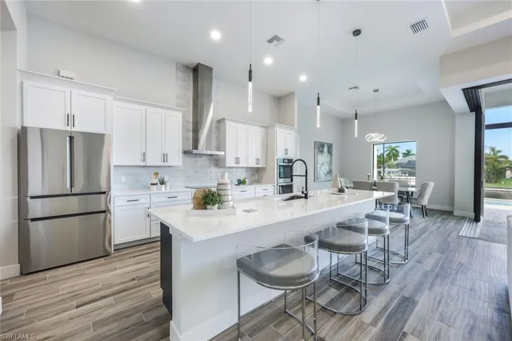 Kitchen featuring appliances with stainless steel finishes, a large island with sink, white cabinets, wall chimney exhaust hood, and decorative light fixtures