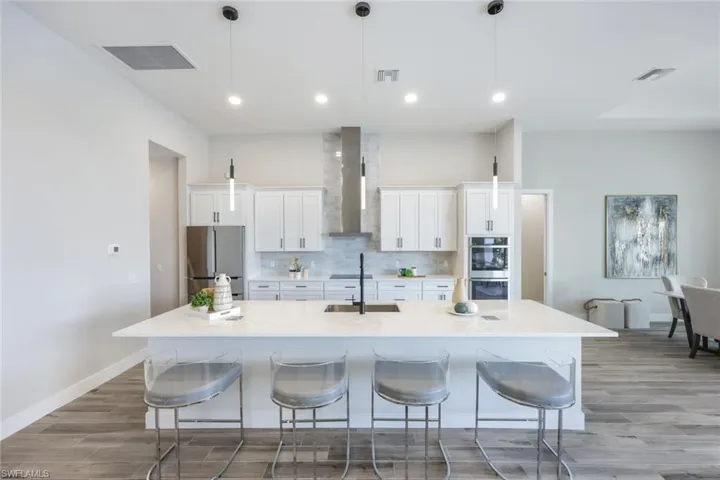 Kitchen with a large island with sink, white cabinets, wall chimney exhaust hood, stainless steel appliances, and decorative light fixtures