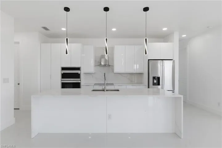 Kitchen featuring light stone countertops, white cabinetry, stainless steel appliances, decorative light fixtures, and an island with sink