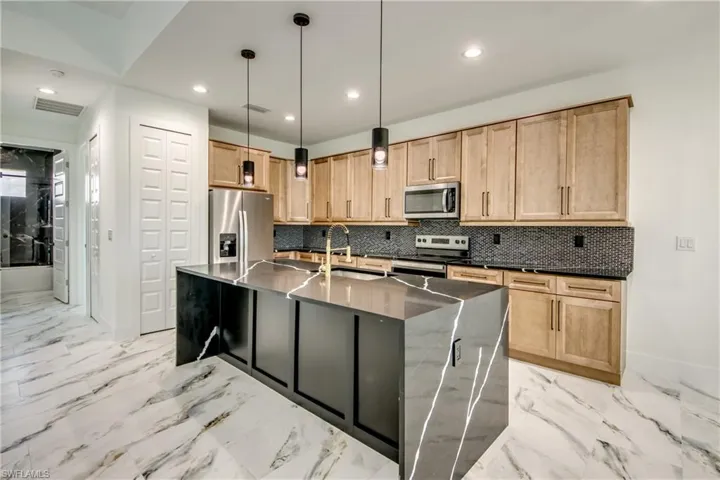 Kitchen featuring stainless steel appliances, backsplash, a center island with sink, hanging light fixtures, and light tile floors