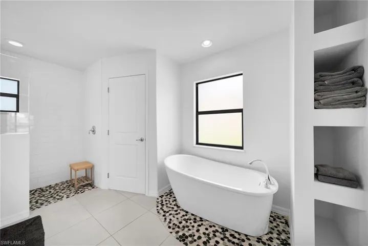 Modern bathroom featuring a freestanding soaking tub, a walk-in shower with pebble tile flooring, built-in recessed shelving, a window with black trim, and recessed lighting
