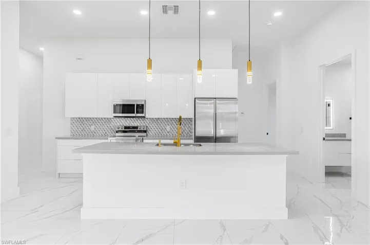 Kitchen featuring light stone countertops, stainless steel appliances, white cabinets, decorative light fixtures, and an island with sink