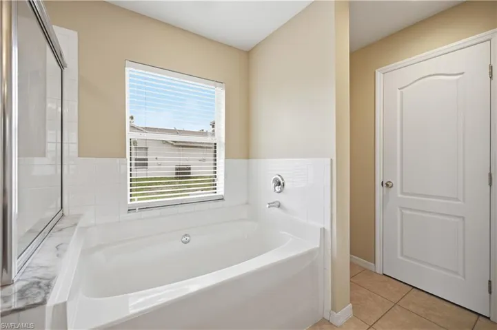 Soaking tub with a white tile surround, chrome fixtures, and a window with horizontal blinds