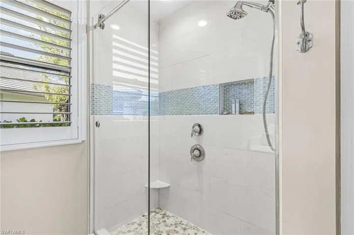 Glass-enclosed shower with white tiled walls, a decorative mosaic tile band, a built-in shower niche, and a contrasting pebble-tiled floor
