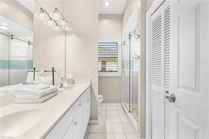 Dual vanity with integrated sinks, frameless glass shower enclosure, louvered bi-fold doors, white cabinetry, and a single window with horizontal blinds