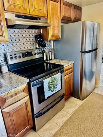 Kitchen with stainless steel appliances, brown cabinets, light tile patterned flooring, under cabinet range hood, and light countertops