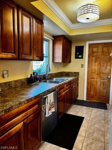 Kitchen featuring crown molding, dark countertops, stainless steel dishwasher, light tile patterned floors, and a tray ceiling