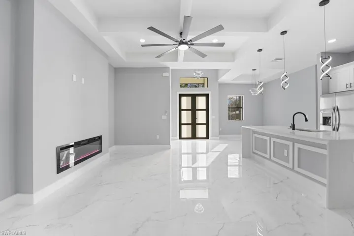 Open-concept interior featuring large-format polished tile flooring, tray ceilings with recessed lighting, a built-in wall-mounted electric fireplace, and a kitchen island with stone-finish countertops and pendant lighting