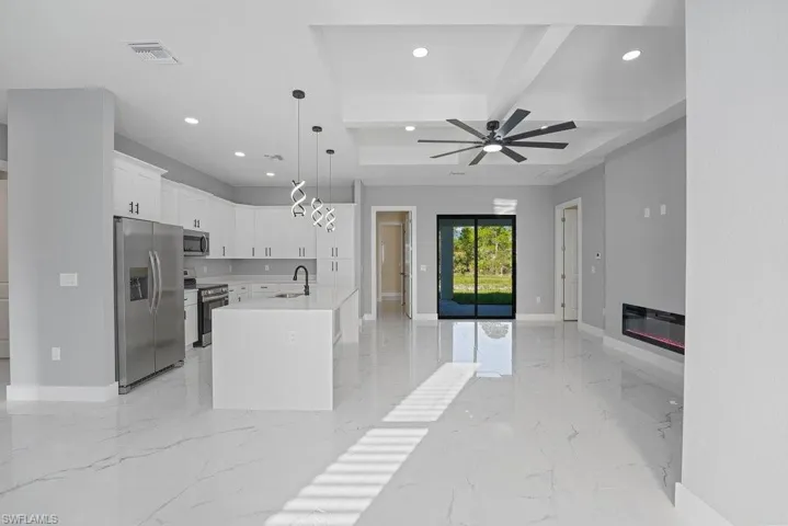 Open-concept interior featuring large-format tile flooring, a chef's kitchen with white shaker cabinetry, quartz countertops, a modern linear electric fireplace, and a tray ceiling with a multi-blade ceiling fan