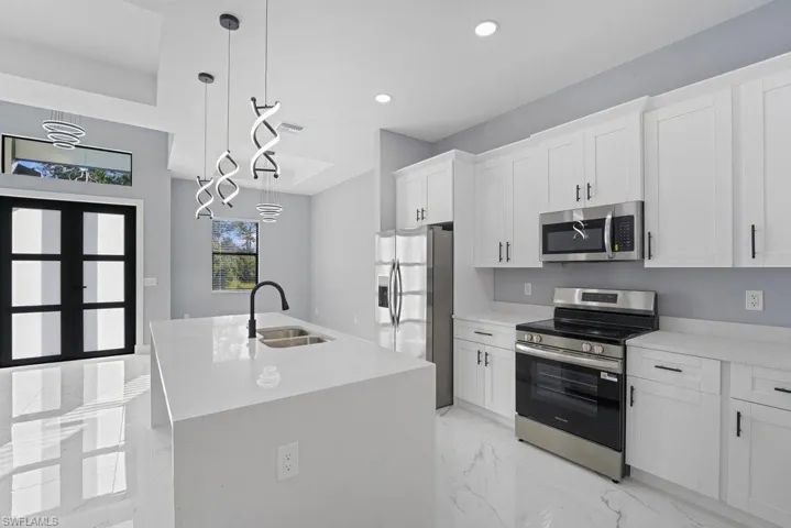Kitchen featuring white cabinetry with matte black hardware, quartz countertops, a central island with a dual-basin undermount sink, stainless steel appliances, and sculptural LED pendant lighting