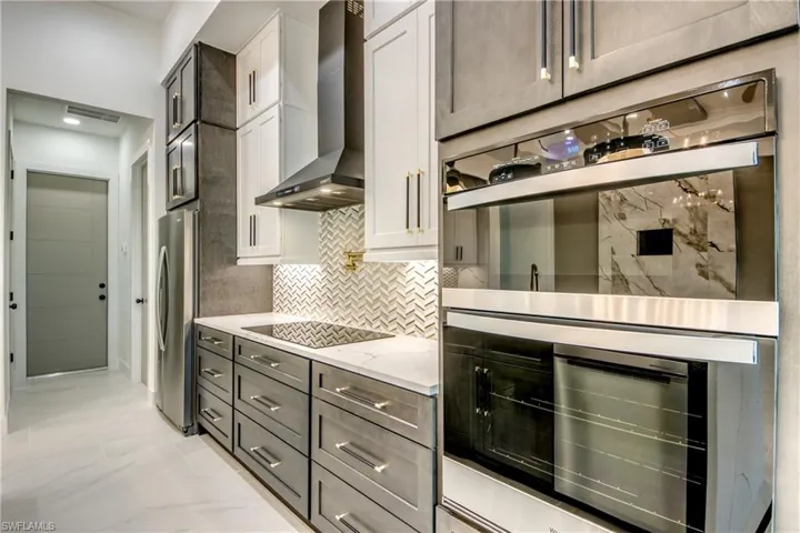 Kitchen with light stone countertops, decorative backsplash, wall chimney range hood, white cabinetry, and appliances with stainless steel finishes