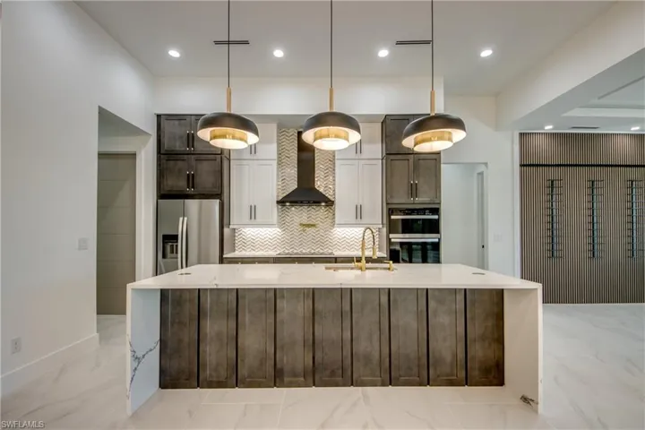 Kitchen featuring a large island with sink, light stone countertops, stainless steel appliances, wall chimney exhaust hood, and pendant lighting