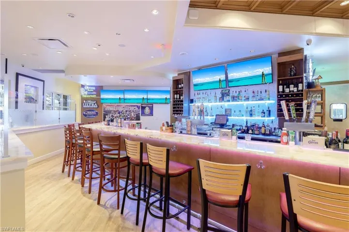 Expansive bar area featuring a light-toned counter surface, built-in shelving, integrated lighting, several wall-mounted display screens, and a grid-patterned ceiling detail