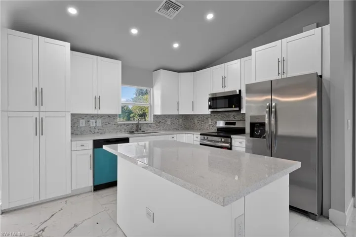 Contemporary kitchen featuring white cabinetry, a large center island with a light-toned countertop, a stainless steel side-by-side refrigerator, a matching microwave, and a mosaic tile backsplash