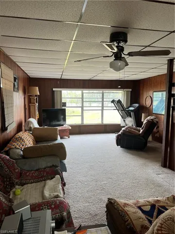 Living room with wooden walls, carpet floors, ceiling fan open to kichen area.