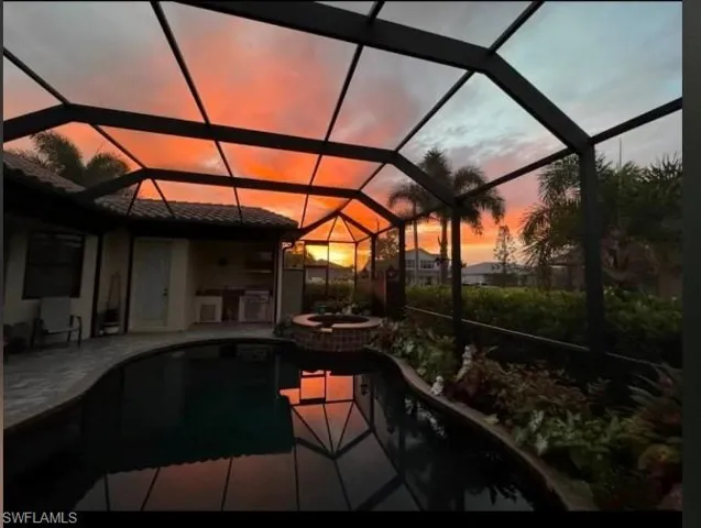 Pool at dusk with exterior kitchen, an in ground hot tub, glass enclosure, and a patio