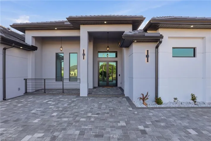Modern exterior with a paver driveway, dark-framed entry doors, vertical accent windows, horizontal railing, and integrated exterior lighting