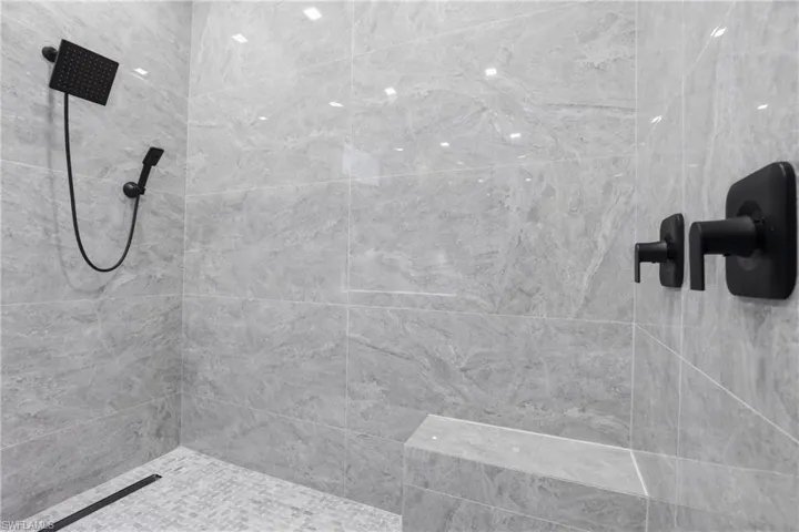 Walk-in shower with large-format grey stone-look tile walls, matte black fixtures, a rain shower head, a handheld sprayer, and a built-in bench