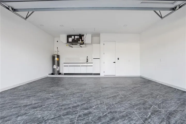 Spacious garage featuring large format dark grey tile flooring, white cabinetry with integrated sink, and a modern white door with black hardware