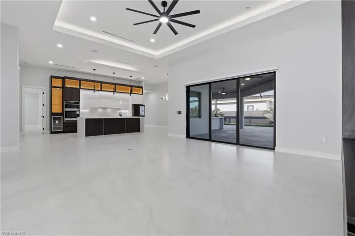 Spacious open-concept interior featuring light-toned flooring, a tray ceiling with recessed lighting and a ceiling fan, and a contemporary kitchen with dark wood-finish cabinetry, integrated appliances, and a central island