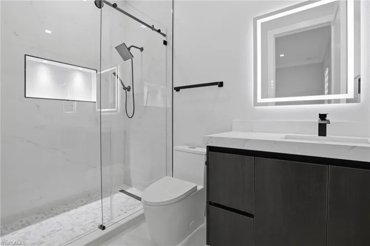 Modern bathroom featuring a frameless glass shower enclosure with matte black fixtures, a built-in shower niche with recessed lighting, a floating vanity with dark wood-finish cabinetry, a white countertop, and an illuminated mirror