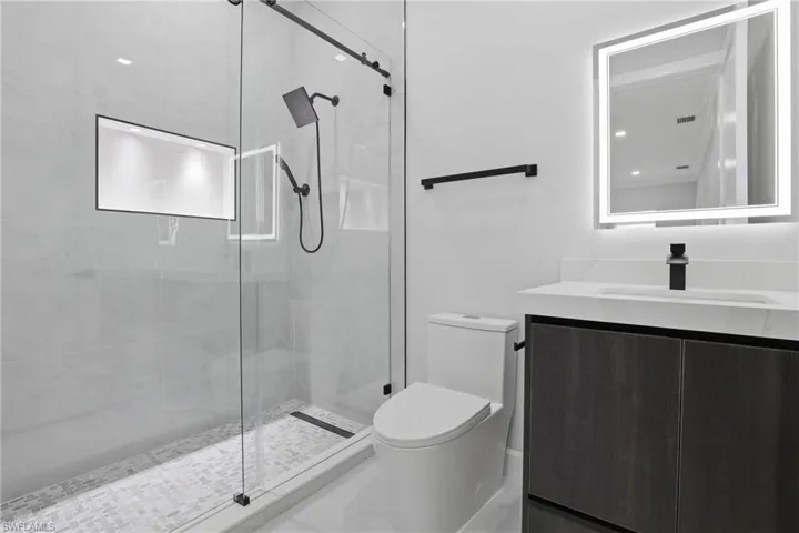 Modern bathroom featuring a frameless glass shower enclosure with matte black hardware, a recessed shower niche with integrated lighting, a dark wood-finish vanity with a white countertop, and an illuminated mirror