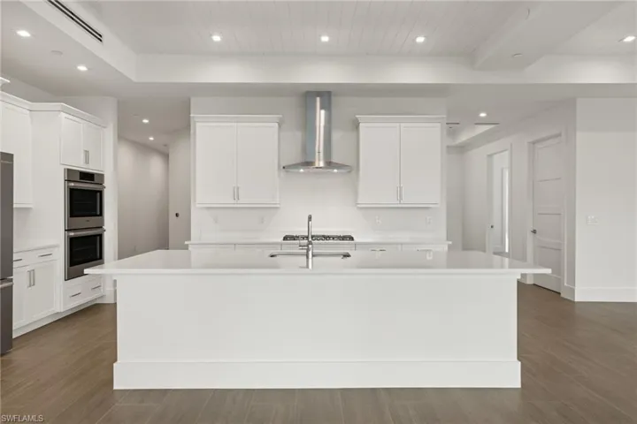 Kitchen featuring Bosch double oven, wall chimney range hood, porcelain tile floors, Quartz island with sink, and white cabinets