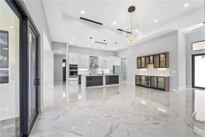 Kitchen with double oven, white cabinetry, hanging light fixtures, a large island with sink, and wall chimney range hood