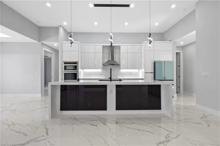 Kitchen featuring double oven, wall chimney range hood, hanging light fixtures, and white cabinets