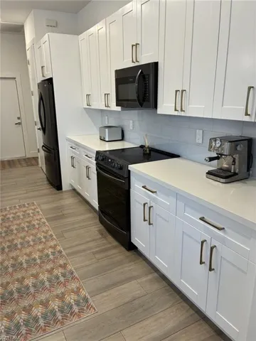 Kitchen featuring white shaker-style cabinetry, light-toned countertops, a light blue subway tile backsplash, black appliances, and wood-finish flooring