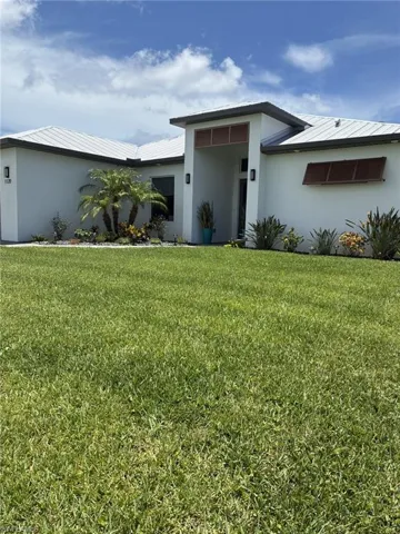Modern stucco exterior featuring a metal roof, prominent entry portico, decorative louvered window coverings, and established landscaping
