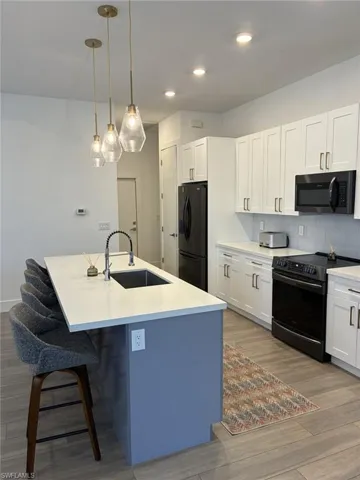 Modern kitchen featuring a spacious island with a built-in sink and pendant lighting, white cabinetry, black appliances, and wood-finish flooring