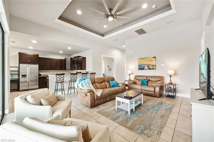 Open-concept living space featuring neutral tile flooring, a tray ceiling with recessed lighting and ceiling fan, and a kitchen with dark wood cabinetry and stainless steel appliances