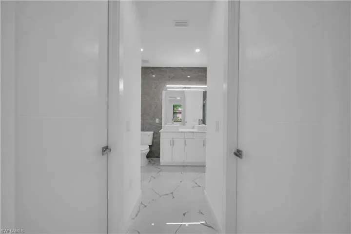 Hallway with white interior doors opening to a bathroom featuring a dual vanity, white cabinetry, and a toilet