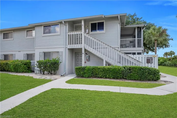 Two-story multi-unit structure featuring horizontal siding, exterior staircase with wood railings, mature landscaping, and a screened balcony