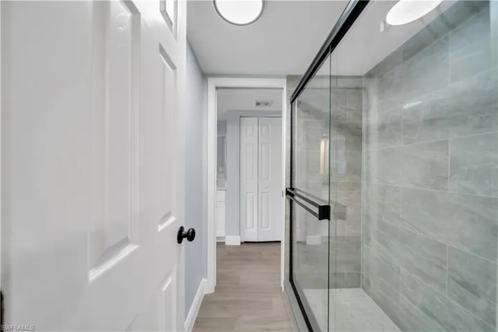 Glass-enclosed shower with matte black framing, textured gray wall tile, light wood-finish flooring, and white paneled doors