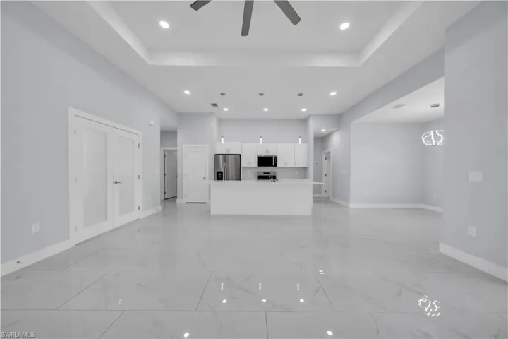 Spacious interior featuring large format tile flooring, a recessed ceiling with integrated lighting, a ceiling fan, and a modern kitchen with white cabinetry, stainless steel appliances, and a central island