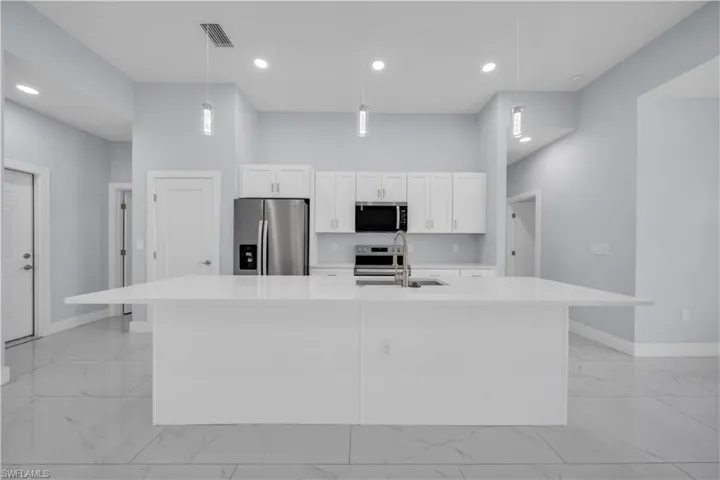 Central kitchen island with a double basin sink, stainless steel appliances, white cabinetry, recessed lighting, and large format tile flooring