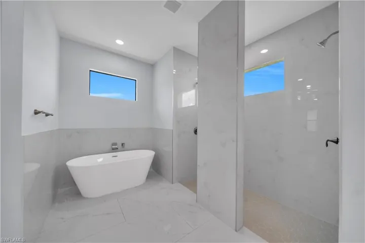 Bathroom featuring a freestanding soaking tub with wall-mounted fixtures, two independent walk-in shower areas, marble-look wall and floor tiling, and rectangular transom windows
