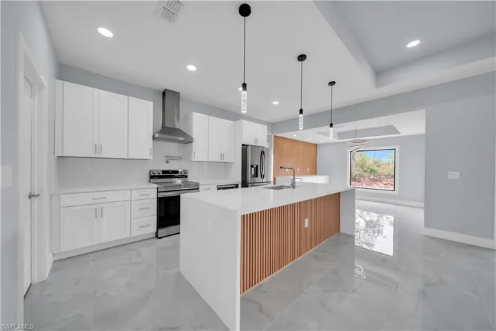 Kitchen with hanging light fixtures, stainless steel appliances, white cabinetry, an island with sink, and light stone countertops