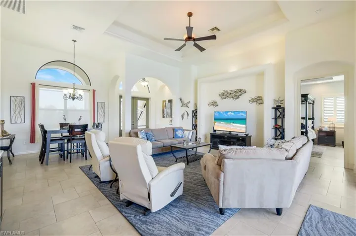 Living room with light tile patterned floors, ceiling fan with notable chandelier, and a raised ceiling